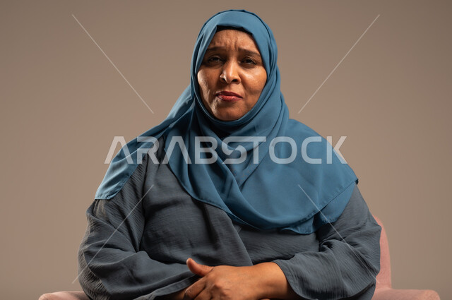  التأمل والتمعن والاندماج, بورتريه لإمرأة كبيرة بالسن عربية خليجية سعودية محجبة ترتدي العباءة الكحلية تجلس على الكرسي بايماءات وجه ويدين مختلفة، الثقة بالنفس والراحة النفسية، خلفية بيج