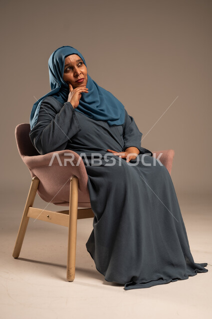  التأمل والتمعن والاندماج, بورتريه لإمرأة كبيرة بالسن عربية خليجية سعودية محجبة ترتدي العباءة الكحلية تجلس على الكرسي بايماءات وجه ويدين مختلفة، الثقة بالنفس والراحة النفسية، خلفية بيج