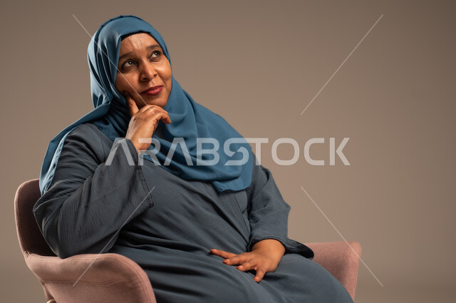  التأمل والتمعن والاندماج, بورتريه لإمرأة كبيرة بالسن عربية خليجية سعودية محجبة ترتدي العباءة الكحلية تجلس على الكرسي بايماءات وجه ويدين مختلفة، الثقة بالنفس والراحة النفسية، خلفية بيج