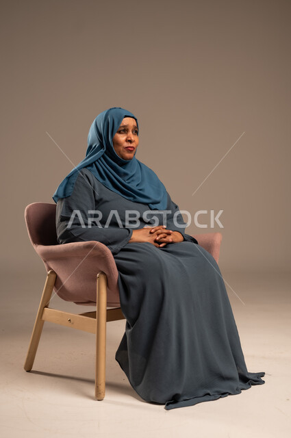  التأمل والتمعن والاندماج, بورتريه لإمرأة كبيرة بالسن عربية خليجية سعودية محجبة ترتدي العباءة الكحلية تجلس على الكرسي بايماءات وجه ويدين مختلفة، الثقة بالنفس والراحة النفسية، خلفية بيج