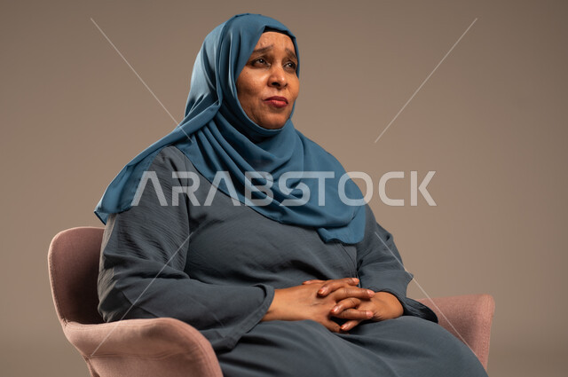  التأمل والتمعن والاندماج, بورتريه لإمرأة كبيرة بالسن عربية خليجية سعودية محجبة ترتدي العباءة الكحلية تجلس على الكرسي بايماءات وجه ويدين مختلفة، الثقة بالنفس والراحة النفسية، خلفية بيج