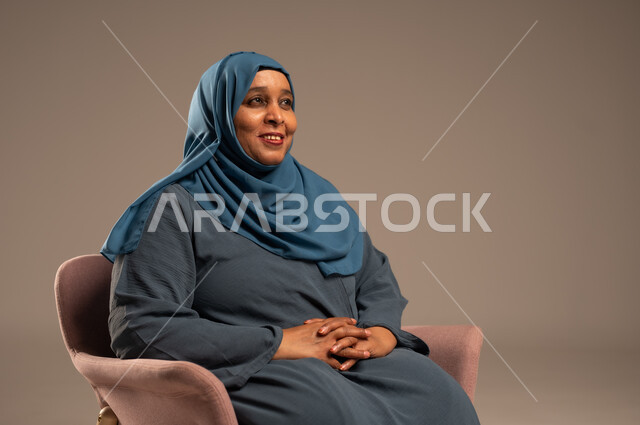 السرور و التأمل والتمعن , بورتريه لإمرأة كبيرة بالسن عربية خليجية سعودية محجبة ترتدي العباءة الكحلية تجلس على الكرسي بايماءات وجه ويدين مختلفة، الثقة بالنفس والراحة النفسية، خلفية بيج