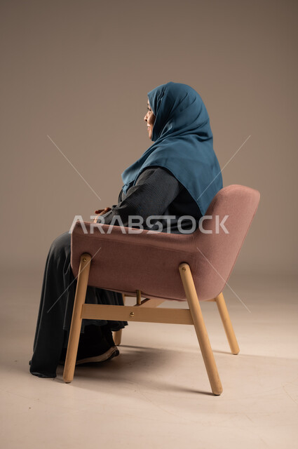 التمعن والتأمل بمكان ما , بورتريه من الجانب لإمرأة كبيرة بالسن عربية خليجية سعودية محجبة ترتدي العباءة الكحلية تجلس على الكرسي بايماءات وجه ويدين مختلفة، الثقة بالنفس والراحة النفسية، خلفية بيج