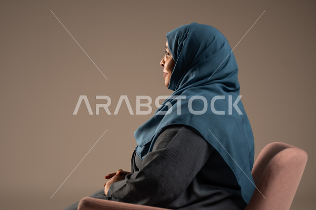 التمعن والتأمل بمكان ما , بورتريه من الجانب لإمرأة كبيرة بالسن عربية خليجية سعودية محجبة ترتدي العباءة الكحلية تجلس على الكرسي بايماءات وجه ويدين مختلفة، الثقة بالنفس والراحة النفسية، خلفية بيج