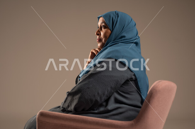 التمعن والتأمل بمكان ما , بورتريه من الجانب لإمرأة كبيرة بالسن عربية خليجية سعودية محجبة ترتدي العباءة الكحلية تجلس على الكرسي بايماءات وجه ويدين مختلفة، الثقة بالنفس والراحة النفسية، خلفية بيج