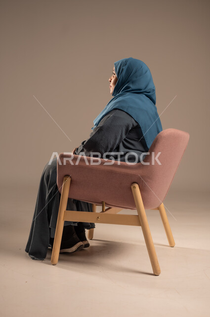 التمعن والتأمل بمكان ما , بورتريه من الجانب لإمرأة كبيرة بالسن عربية خليجية سعودية محجبة ترتدي العباءة الكحلية تجلس على الكرسي بايماءات وجه ويدين مختلفة، الثقة بالنفس والراحة النفسية، خلفية بيج