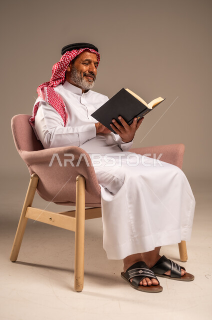 هواية حب المطالعة و الاستكشاف, مفهوم محو الامية، قضاء وقت الفراغ بالتعلم والثقافة، بورتريه لرجل كبير بالسن عربي خليجي سعودي  يرتدي الثوب التقليدي والشماغ يحمل بيده كتاب علمي تثقيفي، ايماءات السعادة والسرور، خلفية بيج