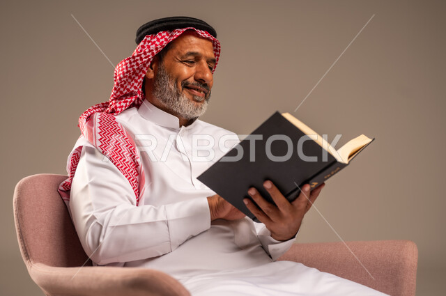 هواية حب المطالعة و الاستكشاف, مفهوم محو الامية، قضاء وقت الفراغ بالتعلم والثقافة، بورتريه لرجل كبير بالسن عربي خليجي سعودي  يرتدي الثوب التقليدي والشماغ يحمل بيده كتاب علمي تثقيفي، ايماءات السعادة والسرور، خلفية بيج