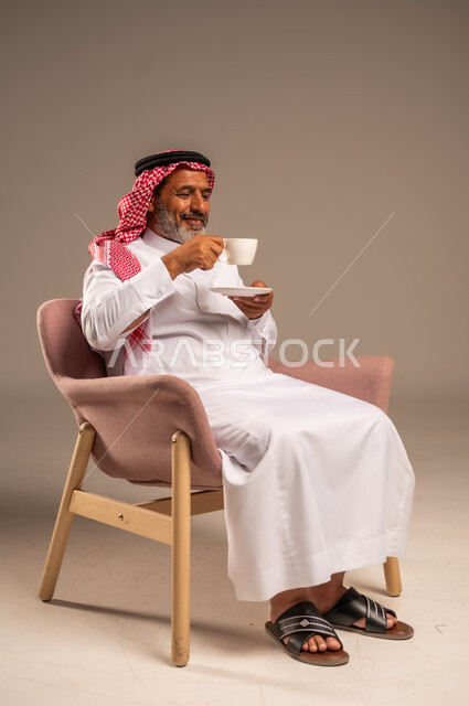 تعديل المزاج بشرب الكافيئين، الاستمتاع بتناول المشروبات الساخنة، بورتريه لرجل مسن عربي خليجي سعودي يرتدي الشماغ والثوب التقليدي يجلس على كرسي ويحمل كوب من القهوة، ايماءات السعادة والسرور، خلفية بيج