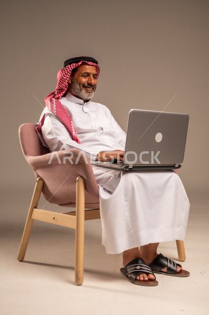،تصفح برامج التواصل الاجتماعي, عقد إجتماع عمل عبر الإنترنت، إستخدام الحاسوب المحمول لإنجاز المهام، بورتريه لرجل عربي خليجي سعودي يرتدي الثوب التقليدي والشماغ يحمل جهاز اللابتوب يجلس على كرسي مريح، تطور التكنولوجيا والأجهزة التقنية، خلفية بيج