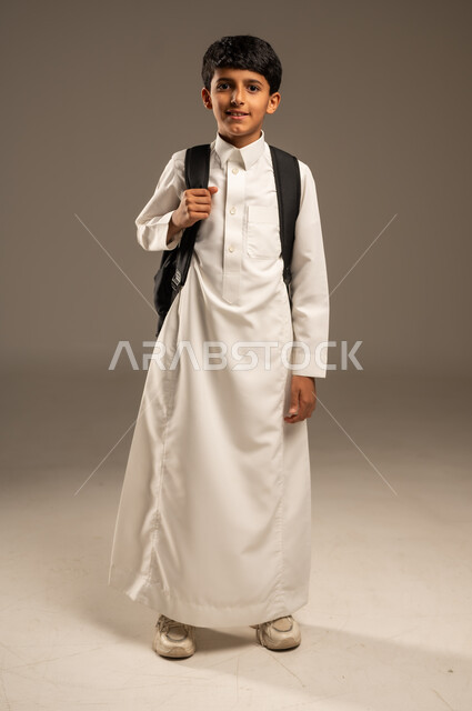 موسم العودة إلى المدارس، التعليم الأكاديمي في المؤسسات التعليمية بالمملكة العربية السعودية، بورتريه لطالب عربي خليجي سعودي يرتدي الثوب التقليدي يحمل حقيبة الظهر المدرسية وينظر إلى الكاميرا بإيماءات السرور والبشاشة، الجسم بالطول الكامل، خلفية رمادية
