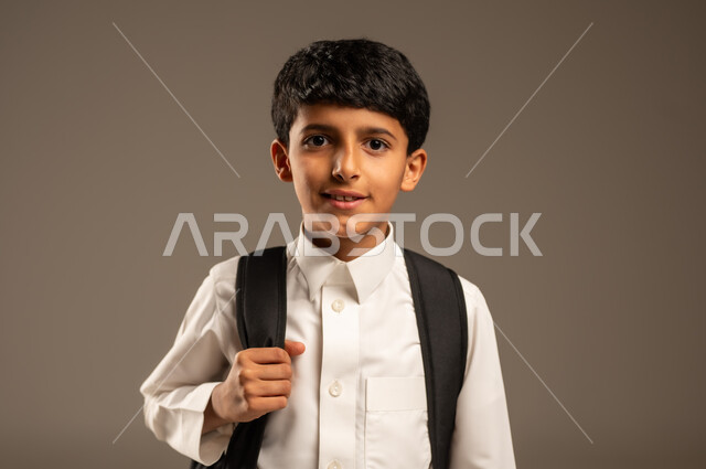 الاستعداد والتجهيز لموسم العودة إلى المدارس، التعليم الأكاديمي في المؤسسات التعليمية بالمملكة العربية السعودية، بورتريه مقرب لطالب عربي خليجي سعودي يرتدي الثوب التقليدي يحمل حقيبة الظهر المدرسية وينظر إلى الكاميرا بإيماءات السرور والبشاشة، خلفية رمادية
