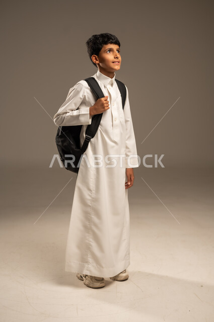  التعليم الأكاديمي في المؤسسات التعليمية بالمملكة العربية السعودية، الاستعداد للذهاب إلى المدرسة، بورتريه من الجانب لطالب عربي خليجي سعودي يرتدي الثوب التقليدي يحمل حقيبة الظهر المدرسية وينظر إلى مكان ما بإيماءات الأمل، الجسم بالطول الكامل، خلفية رمادية
