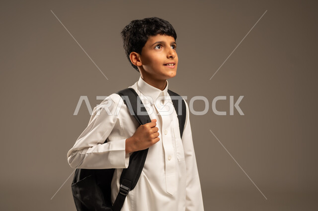 الاستعداد للذهاب إلى المدرسة، موسم العودة إلى المدارس، التعليم الأكاديمي في المؤسسات التعليمية بالمملكة العربية السعودية، بورتريه من الجانب لطالب عربي خليجي سعودي يرتدي الثوب التقليدي يحمل حقيبة الظهر المدرسية وينظر إلى مكان ما بإيماءات الأمل، خلفية رمادية