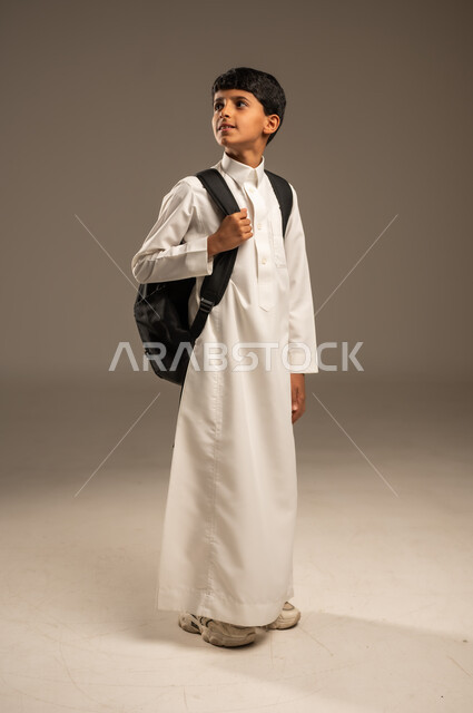  التعليم الأكاديمي في المؤسسات التعليمية بالمملكة العربية السعودية، الاستعداد للذهاب إلى المدرسة، بورتريه من الجانب لطالب عربي خليجي سعودي يرتدي الثوب التقليدي يحمل حقيبة الظهر المدرسية وينظر إلى مكان ما بإيماءات الأمل، الجسم بالطول الكامل، خلفية رمادية