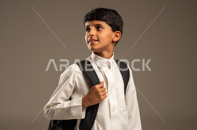 الاستعداد للذهاب إلى المدرسة، موسم العودة إلى المدارس، التعليم الأكاديمي في المؤسسات التعليمية بالمملكة العربية السعودية، بورتريه من الجانب لطالب عربي خليجي سعودي يرتدي الثوب التقليدي يحمل حقيبة الظهر المدرسية وينظر إلى مكان ما بإيماءات الأمل، خلفية رمادية