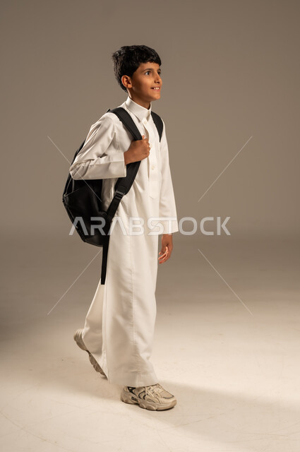 التعليم الأكاديمي في المؤسسات التعليمية بالمملكة العربية السعودية، الاستعداد للذهاب إلى المدرسة، بورتريه من الجانب لطالب عربي خليجي سعودي يرتدي الثوب التقليدي يحمل حقيبة الظهر المدرسية وينظر إلى مكان ما بإيماءات الأمل، الجسم بالطول الكامل، خلفية رمادية