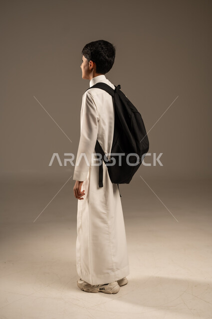  التعليم الأكاديمي في المؤسسات التعليمية بالمملكة العربية السعودية، الاستعداد للذهاب إلى المدرسة، بورتريه من الجانب لطالب عربي خليجي سعودي يرتدي الثوب التقليدي يحمل حقيبة الظهر المدرسية وينظر إلى مكان ما بإيماءات الأمل، الجسم بالطول الكامل، خلفية رمادية
