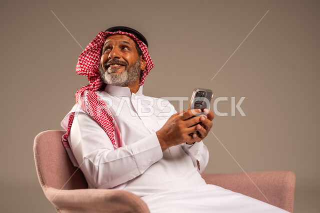 النظر الى شيء ما بايماءات السعادة والسرور، حوارات ودردشات مع الاهل والاقارب، بث مباشر عبر الانترنت، استخدام التقنيات الحديثة والتكنولوجيا، بورتريه لرجل مسن عربي سعودي خليجي يرتدي الثوب التقليدي والشماغ  يجلس على كرسي يحمل بيده الموبايل، خلفية بيج