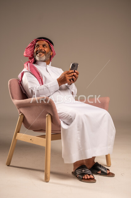 النظر الى شيء ما بايماءات السعادة والسرور، حوارات ودردشات مع الاهل والاقارب، بث مباشر عبر الانترنت، استخدام التقنيات الحديثة والتكنولوجيا، بورتريه لرجل مسن عربي سعودي خليجي يرتدي الثوب التقليدي والشماغ  يجلس على كرسي يحمل بيده الموبايل، خلفية بيج