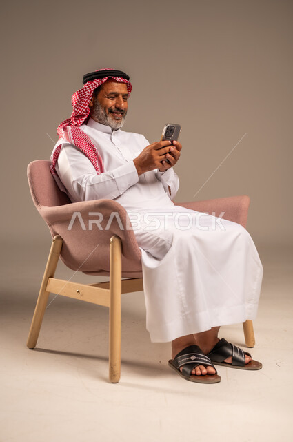 حوارات ودردشات مع الاهل والاقارب، ايماءات السعادة والسرور، بث مباشر عبر الانترنت، استخدام التقنيات الحديثة والتكنولوجيا، بورتريه لرجل مسن عربي سعودي خليجي يرتدي الثوب التقليدي والشماغ  يجلس على كرسي ويحمل بيده الموبايل، خلفية بيج