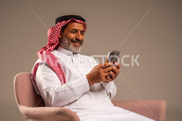حوارات ودردشات مع الاهل والاقارب، ايماءات السعادة والسرور، بث مباشر عبر الانترنت، استخدام التقنيات الحديثة والتكنولوجيا، بورتريه لرجل مسن عربي سعودي خليجي يرتدي الثوب التقليدي والشماغ  يجلس على كرسي ويحمل بيده الموبايل، خلفية بيج