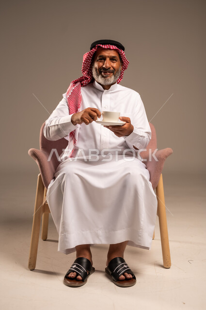 تعديل المزاج بشرب الكافيئين، الاستمتاع بتناول المشروبات الساخنة، بورتريه لرجل مسن عربي خليجي سعودي يرتدي الشماغ والثوب التقليدي يجلس على كرسي ويحمل كوب من القهوة ينظر الى بايماءات السعادة والسرور، خلفية بيج