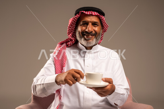 تعديل المزاج بشرب الكافيئين، الاستمتاع بتناول المشروبات الساخنة، بورتريه مقرب لرجل مسن عربي خليجي سعودي يرتدي الشماغ والثوب التقليدي يجلس على كرسي ويحمل كوب من القهوة ينظر الى بايماءات السعادة والسرور، خلفية بيج