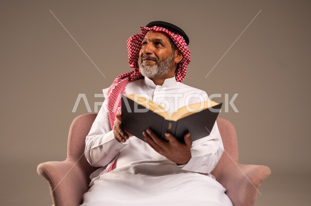 قضاء وقت الفراغ بالتعلم والثقافة، مفهوم محو الامية، هواية حب المطالعة والاستكشاف, بورتريه لرجل كبير بالسن عربي خليجي سعودي  يرتدي الثوب التقليدي والشماغ يجلس على الكرسي يحمل بيده كتاب علمي تثقيفي وينظر الى شيء ما، خلفية بيج