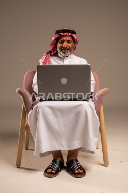 Browsing social media, using laptop to complete tasks, portrait of a Saudi Arabian Gulf man wearing traditional thobe and shemagh holding a laptop sitting on a comfortable chair, development of technology and technical devices, beige background