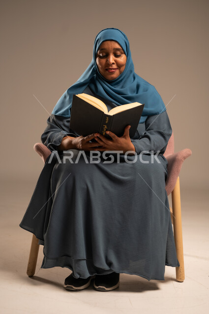 مفهوم محو الامية، هواية حب المطالعة و الاستكشاف, قضاء وقت الفراغ بالتعلم والثقافة، بورتريه مقرب لامراة عربية خليجية سعودية محجبة ترتدي العباءة تجلس على كرسي تحمل بيدها كتاب علمي تثقيفي، خلفية بيج