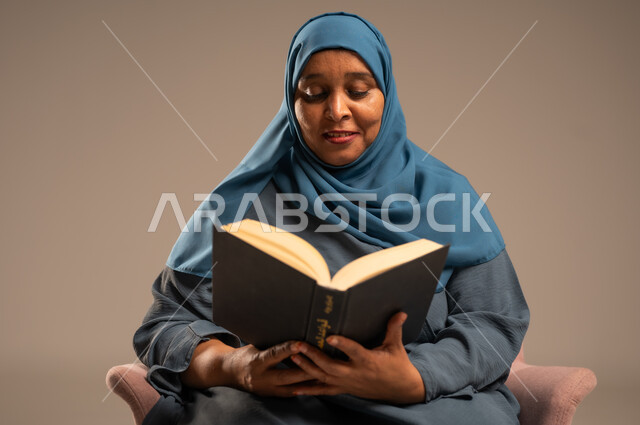 مفهوم محو الامية، هواية حب المطالعة و الاستكشاف, قضاء وقت الفراغ بالتعلم والثقافة، بورتريه مقرب لامراة عربية خليجية سعودية محجبة ترتدي العباءة تجلس على كرسي تحمل بيدها كتاب علمي تثقيفي، خلفية بيج