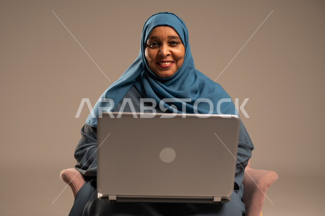 عقد إجتماع عمل عبر الإنترنت، تصفح برامج التواصل الاجتماعي, إستخدام الحاسوب المحمول لإنجاز المهام، بورتريه لامراة عربية خليجية سعودية ترتدي العباءة والحجاب تجلس على كرسي تحمل جهاز اللابتوب، تطور التكنولوجيا والأجهزة التقنية، خلفية بيج