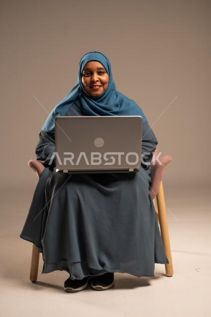 عقد إجتماع عمل عبر الإنترنت، تصفح برامج التواصل الاجتماعي, إستخدام الحاسوب المحمول لإنجاز المهام، بورتريه لامراة عربية خليجية سعودية ترتدي العباءة والحجاب تجلس على كرسي تحمل جهاز اللابتوب، تطور التكنولوجيا والأجهزة التقنية، خلفية بيج