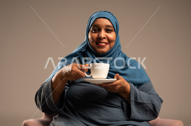 تعديل المزاج بشرب الكافيئين، الاستمتاع بتناول المشروبات الساخنة، بورتريه لامراه عربية خليجية سعودية محجبة ترتدي العباءة تجلس على كرسي وتحمل كوب من القهوة تنظر الى الكاميرا بايماءات السعادة والسرور، خلفية بيج