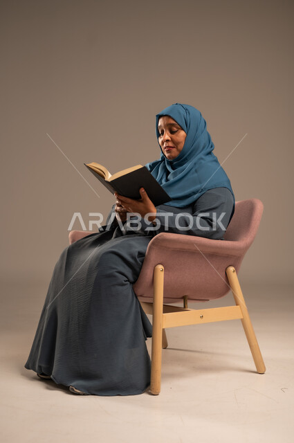 The concept of literacy, the hobby of reading and exploring, spending free time learning and culture, a portrait of a veiled Saudi Arabian Gulf woman wearing a blue abaya holding an educational scientific book in her hand, beige background