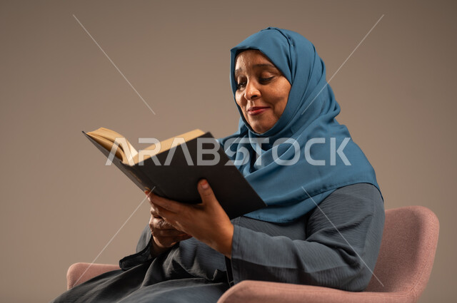 مفهوم محو الامية، هواية حب المطالعة و الاستكشاف, قضاء وقت الفراغ بالتعلم والثقافة، بورتريه لامراة عربية خليجية سعودية محجبة ترتدي العباءة الزرقاء تحمل بيدها كتاب علمي تثقيفي، خلفية بيج