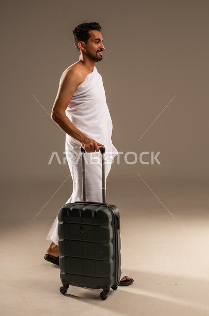 السفر والترحال لزيارة بيت الله الحرام، بورتريه مقرب من الجانب لرجل مسلم خليجي عربي سعودي مبتسم يرتدي ملابس الاحرام يمسك بيده حقيبة سفر سوداء، التقرب الى الله من خلال أداء فريضة الحج والعمرة، النظر الى مكان ما بعيدا  بإيماءات السرور والسعادة، صورة الجسم بالكامل، خلفية بيج