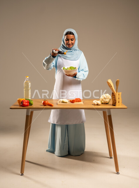  احتراف مهنة الطبخ، المتعة في تحضير الوصفات الشهية, بورتريه لامرأة عربية خليجية سعودية محجبة ترتدي المئزر الأبيض تقف خلف طاولة الطعام الخشبية، إعداد السلطات والخضروات والأطباق الصحية،مهارات تقديم وتزيين الطعام بطرق مبتكرة، الجسم بالطول الكامل، خلفية رمادية