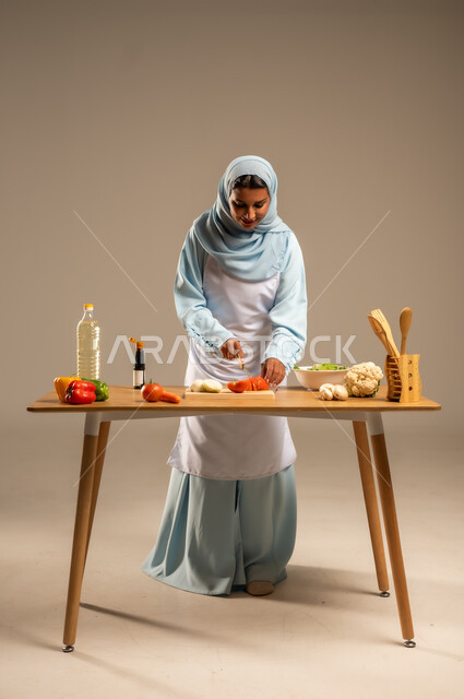  احتراف مهنة الطبخ، المتعة في تحضير الوصفات الشهية, بورتريه لامرأة عربية خليجية سعودية محجبة ترتدي المئزر الأبيض تقف خلف طاولة الطعام الخشبية، إعداد السلطات والخضروات والأطباق الصحية،مهارات تقديم وتزيين الطعام بطرق مبتكرة، الجسم بالطول الكامل، خلفية رمادية