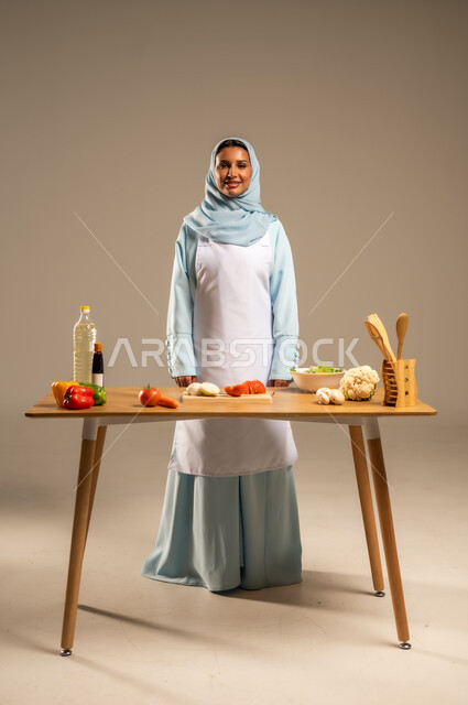  احتراف مهنة الطبخ، المتعة في تحضير الوصفات الشهية, بورتريه لامرأة عربية خليجية سعودية محجبة ترتدي المئزر الأبيض تقف خلف طاولة الطعام الخشبية، إعداد السلطات والخضروات والأطباق الصحية،مهارات تقديم وتزيين الطعام بطرق مبتكرة، الجسم بالطول الكامل، خلفية رمادية
