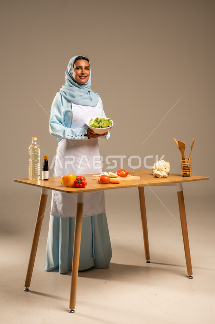  احتراف مهنة الطبخ، المتعة في تحضير الوصفات الشهية, بورتريه لامرأة عربية خليجية سعودية محجبة ترتدي المئزر الأبيض تقف خلف طاولة الطعام الخشبية، إعداد السلطات والخضروات والأطباق الصحية،مهارات تقديم وتزيين الطعام بطرق مبتكرة، الجسم بالطول الكامل، خلفية رمادية