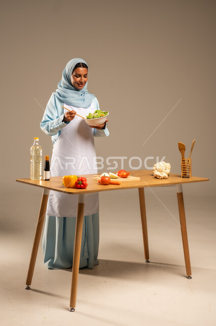  احتراف مهنة الطبخ، المتعة في تحضير الوصفات الشهية, بورتريه لامرأة عربية خليجية سعودية محجبة ترتدي المئزر الأبيض تقف خلف طاولة الطعام الخشبية، إعداد السلطات والخضروات والأطباق الصحية،مهارات تقديم وتزيين الطعام بطرق مبتكرة، الجسم بالطول الكامل، خلفية رمادية