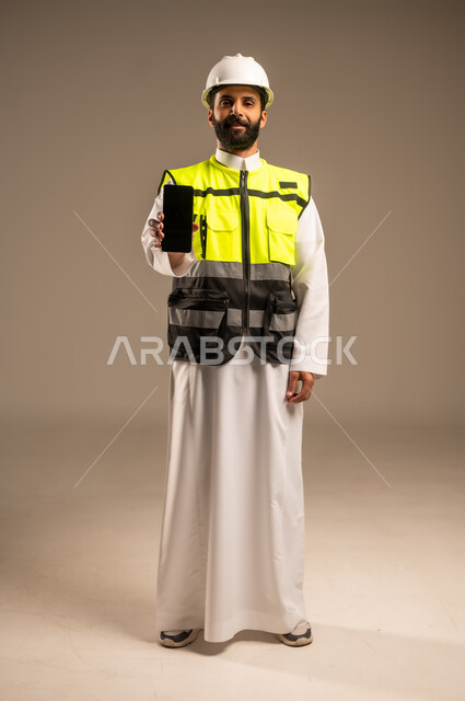عرض شاشة سوداء فارغة، مفهوم الهندسة والانشاءات المعمارية، استخدام الأجهزة التقنية الحديثة، بورتريه لمهندس عربي خليجي سعودي يرتدي الثوب التقليدي والسترة الخاصة وخوذة الحماية يمسك بيده الهاتف المحمول ينظر إلى الكاميرا، الجسم بالطول الكامل، خلفية بيج