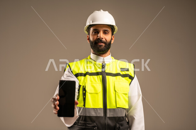 عرض شاشة سوداء فارغة، مفهوم الهندسة والانشاءات المعمارية، استخدام الأجهزة التقنية الحديثة، بورتريه مقرب لمهندس عربي خليجي سعودي يرتدي الثوب التقليدي والسترة الخاصة وخوذة الحماية يمسك بيده الهاتف المحمول ينظر إلى الكاميرا، الإعمار بسواعد أبناء الوطن، خلفية بيج