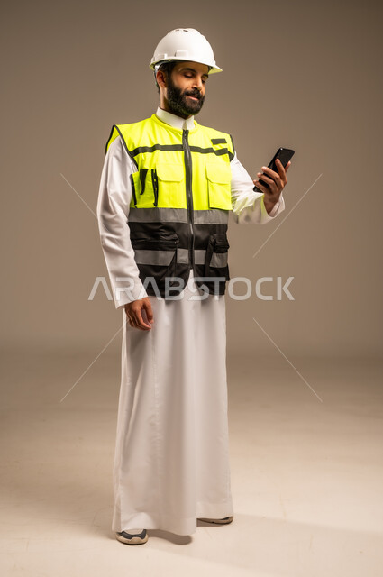 The concept of engineering and architectural construction, the use of modern technical devices, construction by the hands of the sons of the homeland, a portrait of a Saudi Arabian Gulf engineer wearing the traditional dress, a special jacket and a protective helmet, holding a mobile phone in his hand, following up on work, the body in full length, beige background