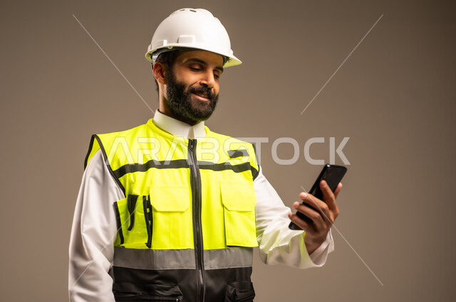 مفهوم الهندسة والانشاءات المعمارية، استخدام الأجهزة التقنية الحديثة، الإعمار بسواعد أبناء الوطن، بورتريه مقرب لمهندس عربي خليجي سعودي يرتدي الثوب التقليدي والسترة الخاصة وخوذة الحماية يمسك بيده الهاتف المحمول يقوم بمتابعة الأعمال، مهن ووظائف سعودية، خلفية بيج