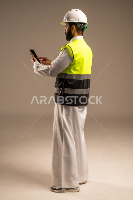 مهن ووظائف سعودية للشباب، استخدام الأجهزة التقنية الحديثة في الهندسة والانشاءات المعمارية، بورتريه من الجانب لمهندس عربي خليجي سعودي يرتدي الثوب التقليدي والسترة الخاصة وخوذة الحماية يمسك بيده الهاتف المحمول يقوم بمتابعة الأعمال، الجسم بالطول الكامل، خلفية بيج