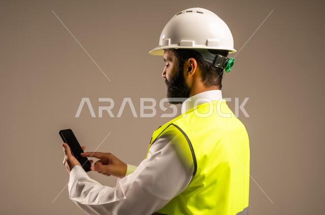 مهن ووظائف سعودية للشباب، استخدام الأجهزة التقنية الحديثة في الهندسة والانشاءات المعمارية، بورتريه مقرب من الجانب لمهندس عربي خليجي سعودي يرتدي الثوب التقليدي والسترة الخاصة وخوذة الحماية يمسك بيده الهاتف المحمول يقوم بمتابعة الأعمال، خلفية بيج