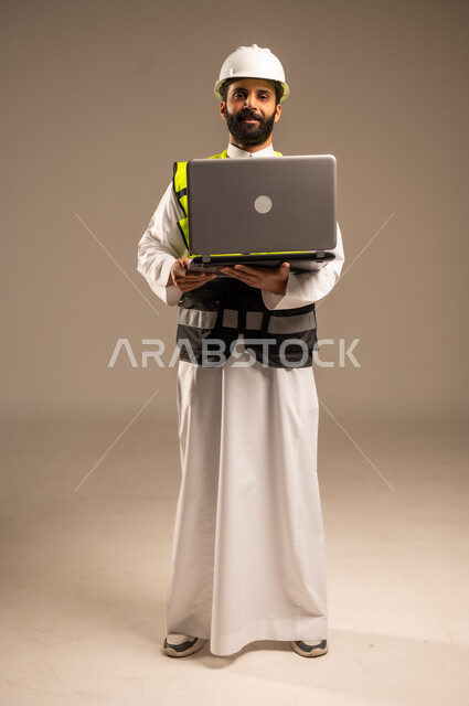 الإعمار بسواعد أبناء الوطن، استخدام أجهزة تقنية حديثة في الهندسة والانشاءات المعمارية، بورتريه لمهندس عربي خليجي سعودي يرتدي الثوب التقليدي والسترة الخاصة وخوذة الحماية يحمل الحاسوب المحمول ينظر إلى الكاميرا يقوم بمتابعة الأعمال، الجسم بالطول الكامل، خلفية بيج